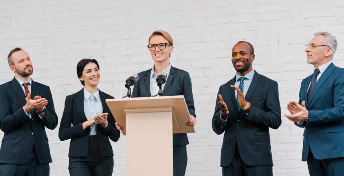 A woman speaking on the mic, other people clapping